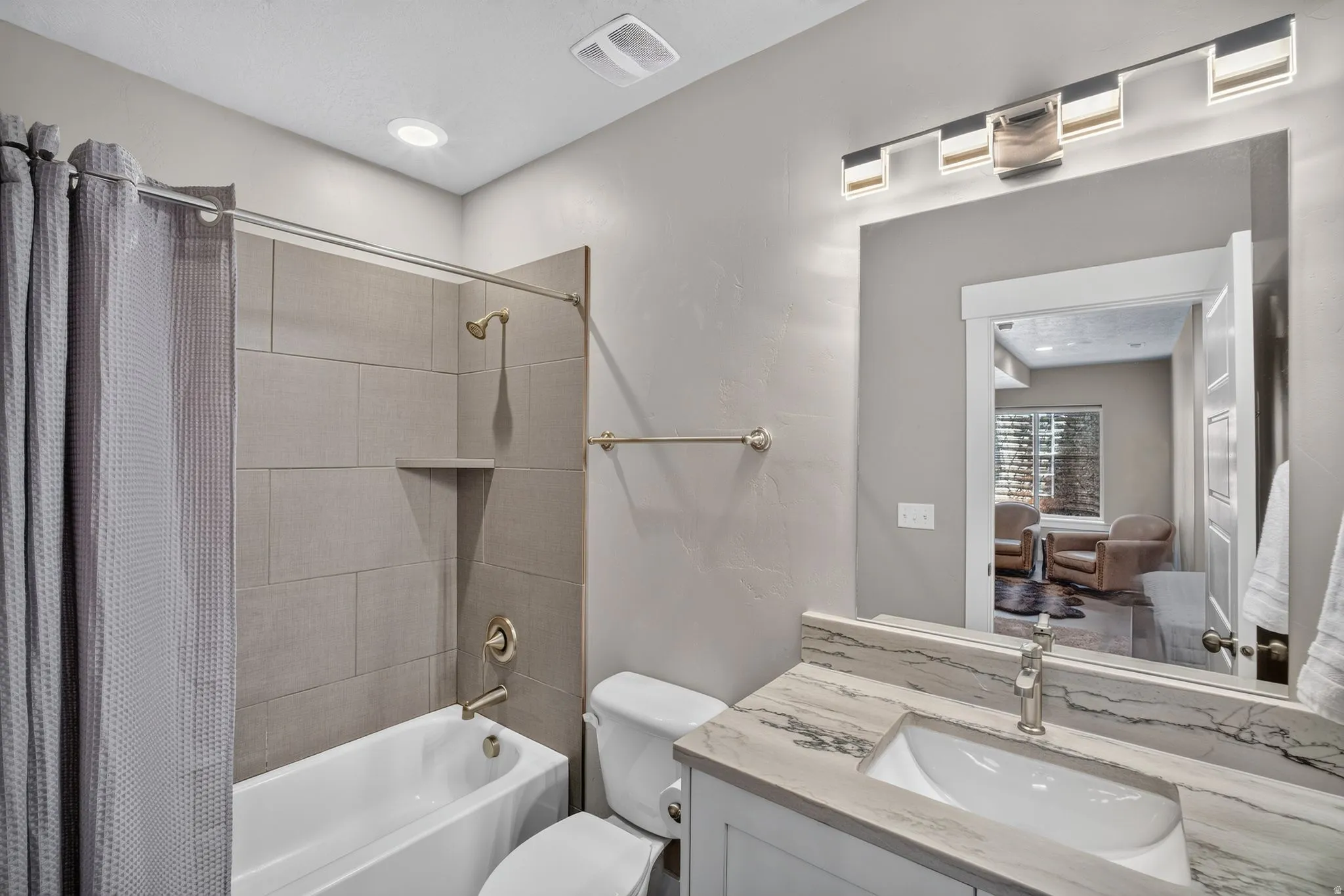 Bathroom featuring a white bathtub with a tiled shower surround, brushed nickel fixtures, a single vanity with a stone countertop, an undermount sink, and a framed mirror with linear lighting