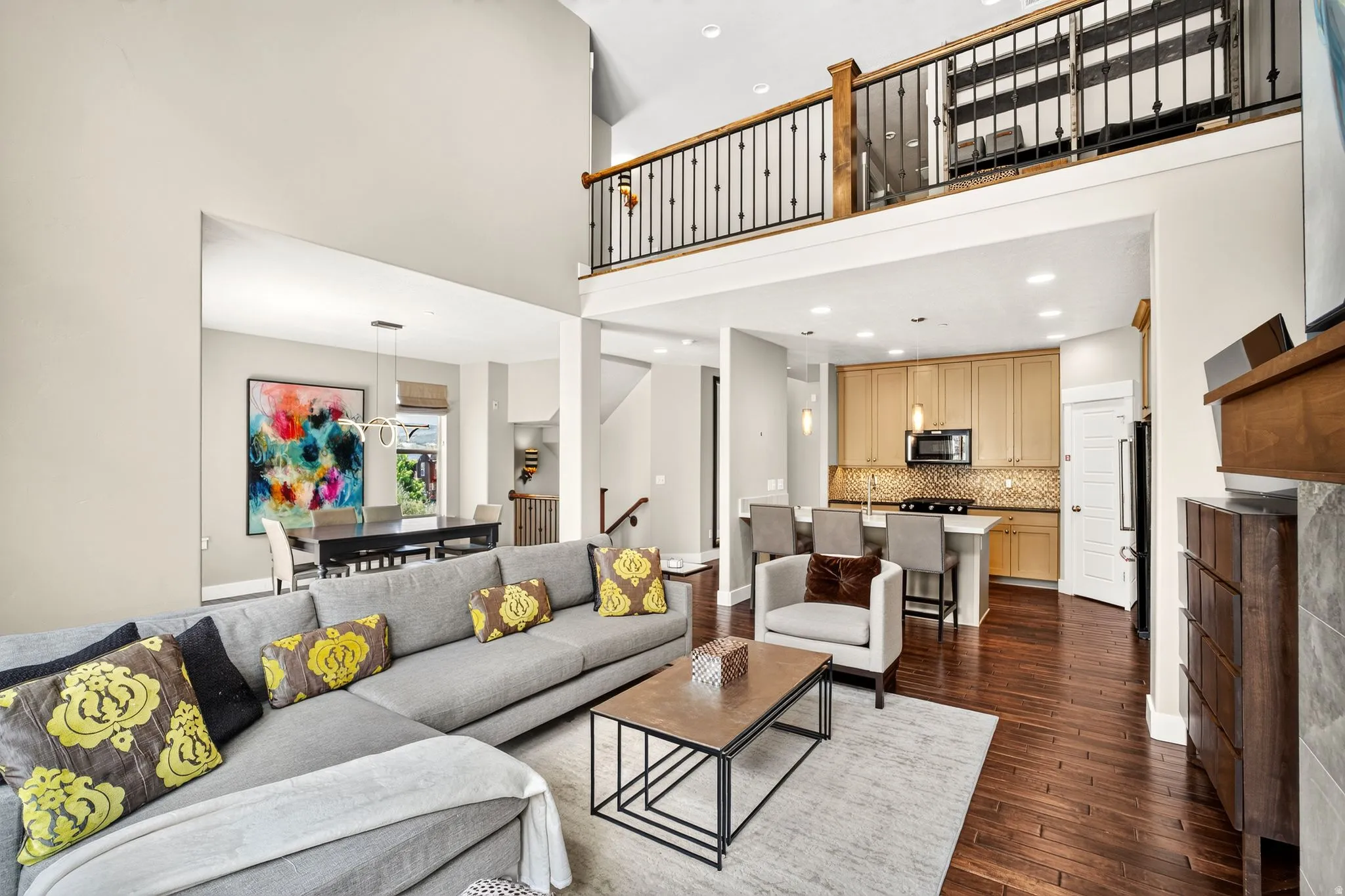 Open-concept living area featuring high ceilings, wood-finish flooring, a kitchen with light-tone cabinetry and stainless steel appliances, a breakfast bar, and an open loft overlooking the space