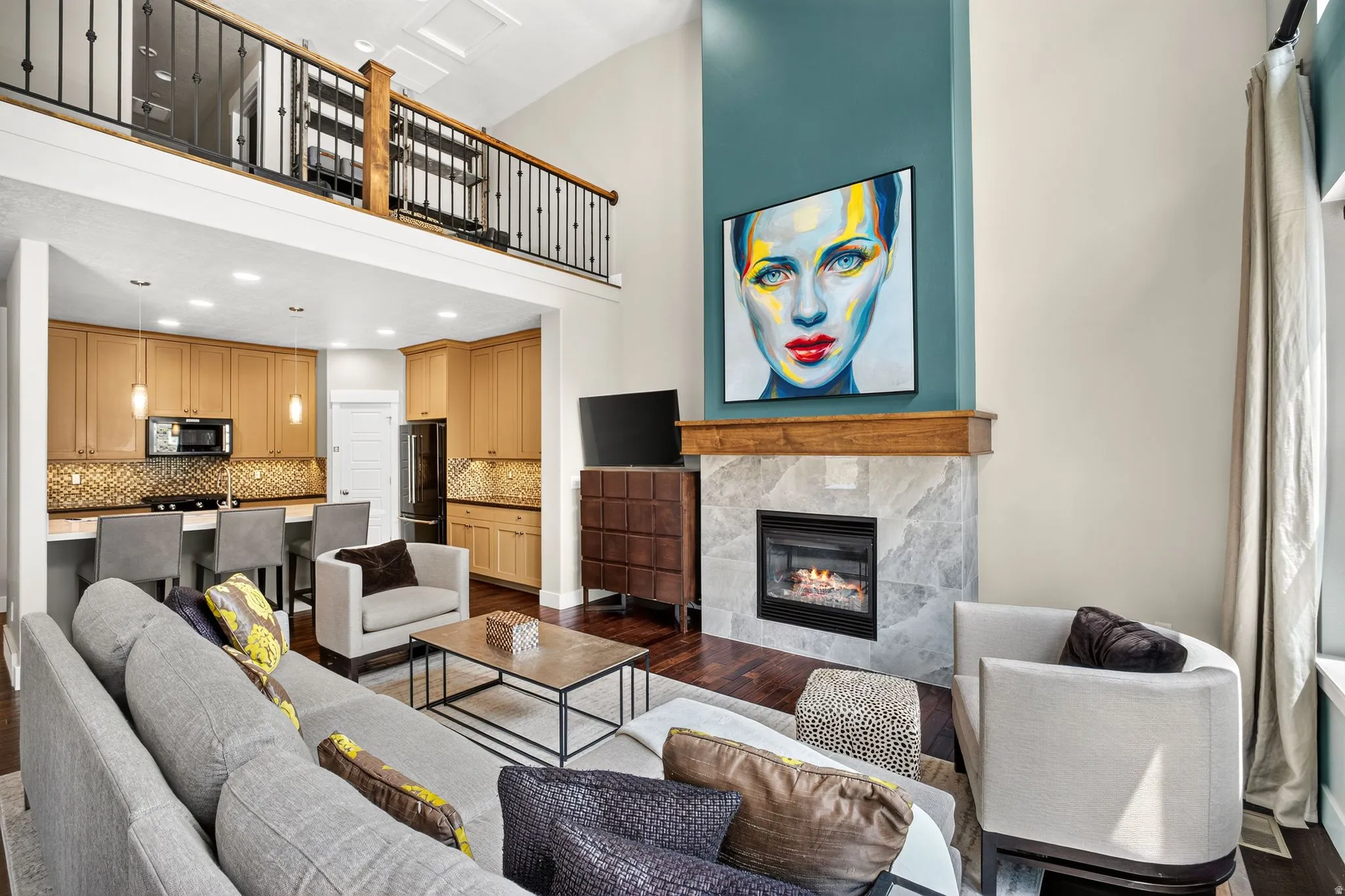 Open-concept living area featuring a double-height ceiling, a fireplace with a stone surround and wood mantel, a loft with an iron railing, wood-finish flooring, and a kitchen with light wood cabinetry and a tiled backsplash