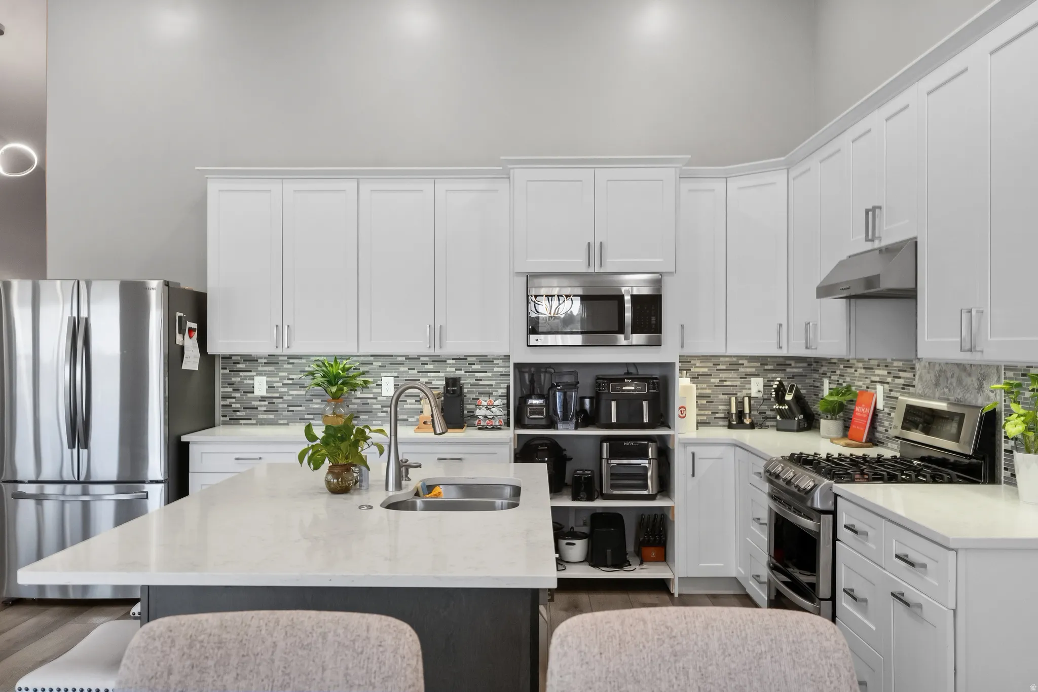 Kitchen featuring an expansive island with a stainless steel undermount sink, white quartz countertops, full-height tile backsplash, white shaker-style cabinetry, and stainless steel appliances