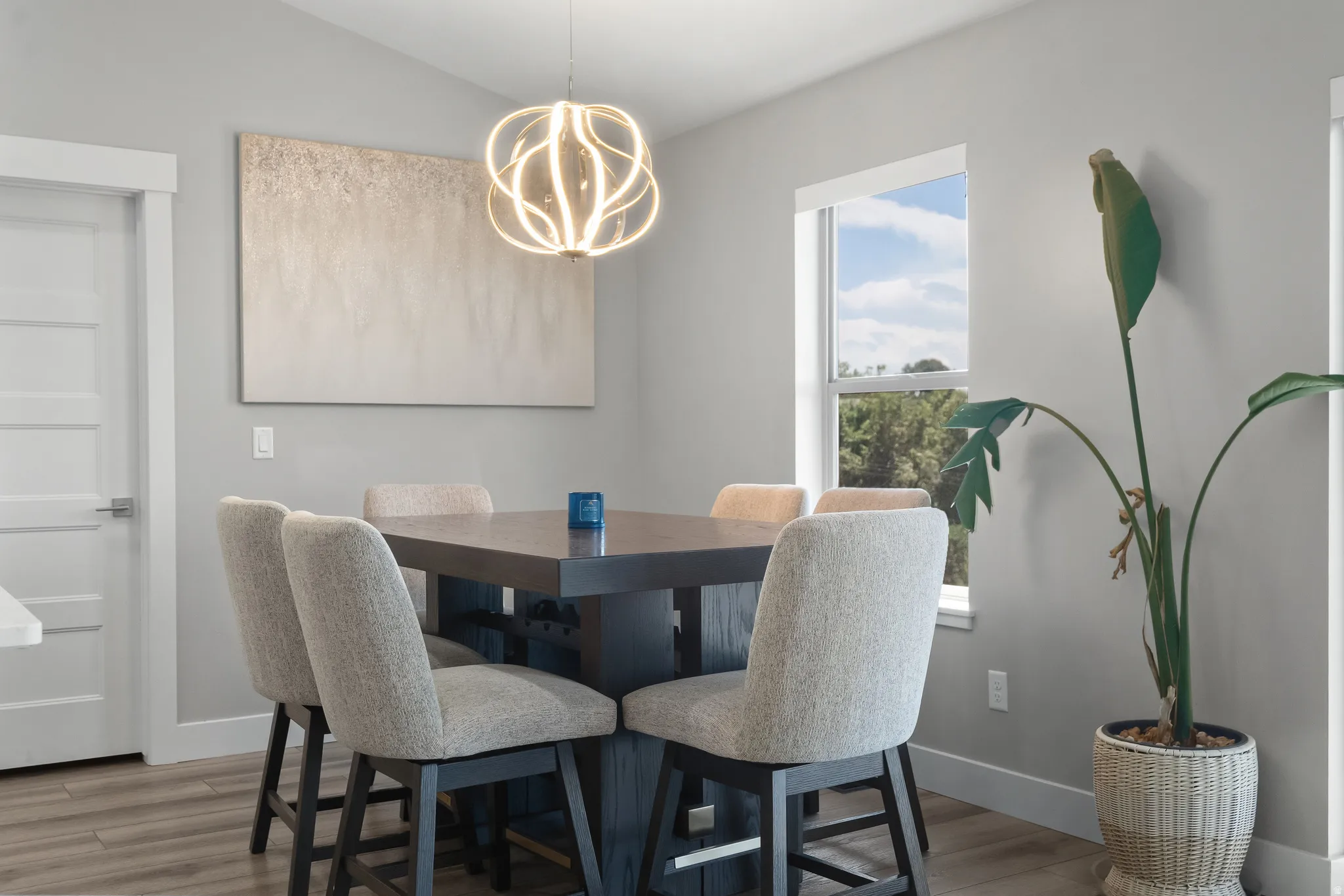 Modern dining space featuring a contemporary chandelier, wood-finish flooring, light grey wall paint, a multi-pane window, and a five-panel interior door