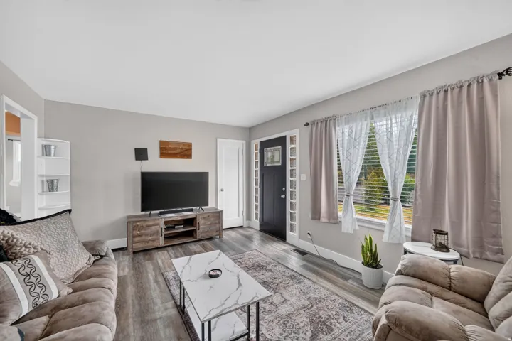 Spacious living area featuring wood-finish flooring, a white trim entry door with sidelights, and large windows with blinds
