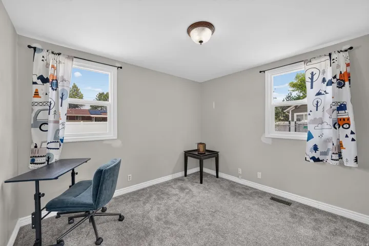 Spacious room featuring light gray walls, plush gray carpeting, and two large windows providing natural illumination
