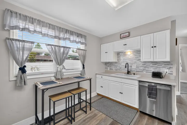 Bright kitchen featuring white cabinetry, a white rectangular sink with a matte black faucet, a textured tile backsplash, stainless steel dishwasher, and wood-finish flooring