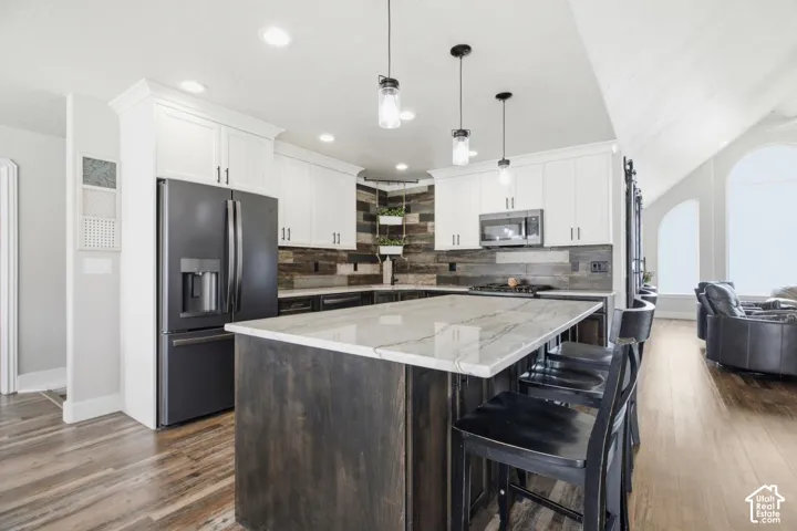 Open-concept kitchen featuring an oversized island with seating, white upper cabinetry, distressed wood-look backsplash, matte black appliances, and pendant lighting