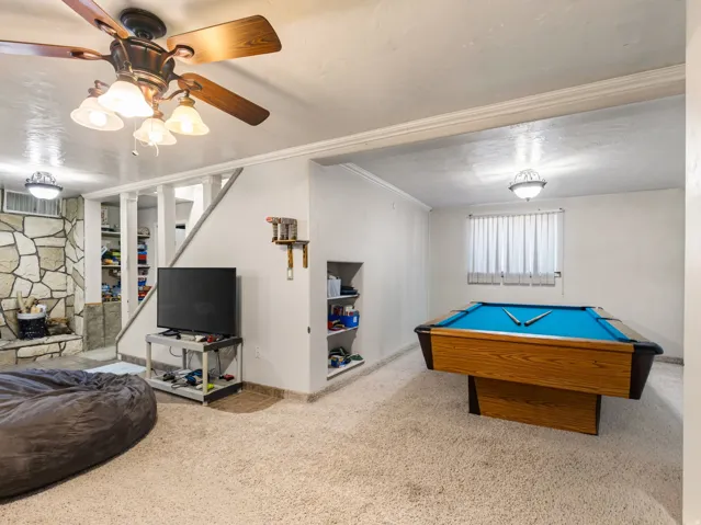 Expansive carpeted room featuring a stone-clad fireplace, integrated shelving units, a ceiling fan with light fixture, and a window with blinds