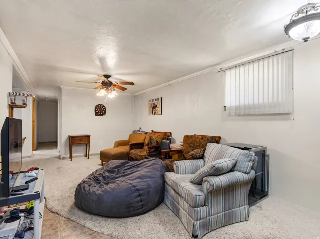 Carpeted living area featuring white walls, crown molding, a ceiling fan with light fixture, and window with vertical blinds