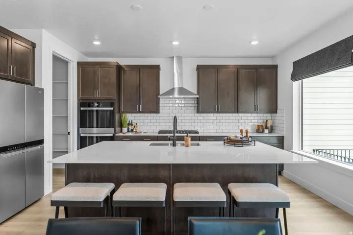 Contemporary kitchen featuring a large central island with seating, stainless steel appliances, dark wood-finish cabinetry, white subway tile backsplash, and a built-in double oven