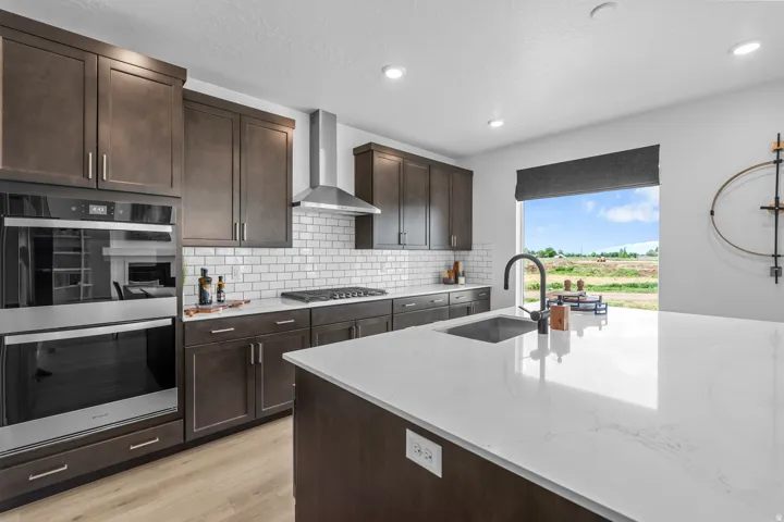 Contemporary kitchen featuring a large island with a composite sink and matte black faucet, dark wood-finish cabinetry, white subway tile backsplash, stainless steel appliances, and wood-finish flooring