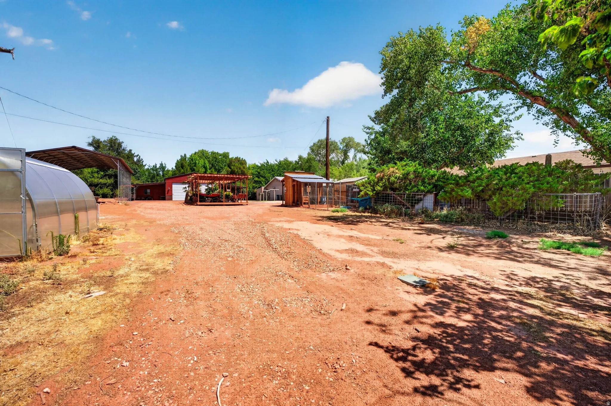 Gravel-surfaced yard featuring a large greenhouse, a covered open-air structure, and a red-painted outbuilding with a roll-up door