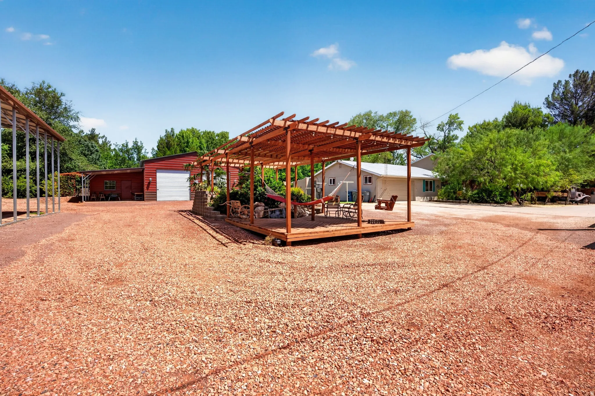 Spacious gravel lot featuring a freestanding wooden pergola, a red metal-sided workshop with a roll-up door, and a light-colored exterior residence