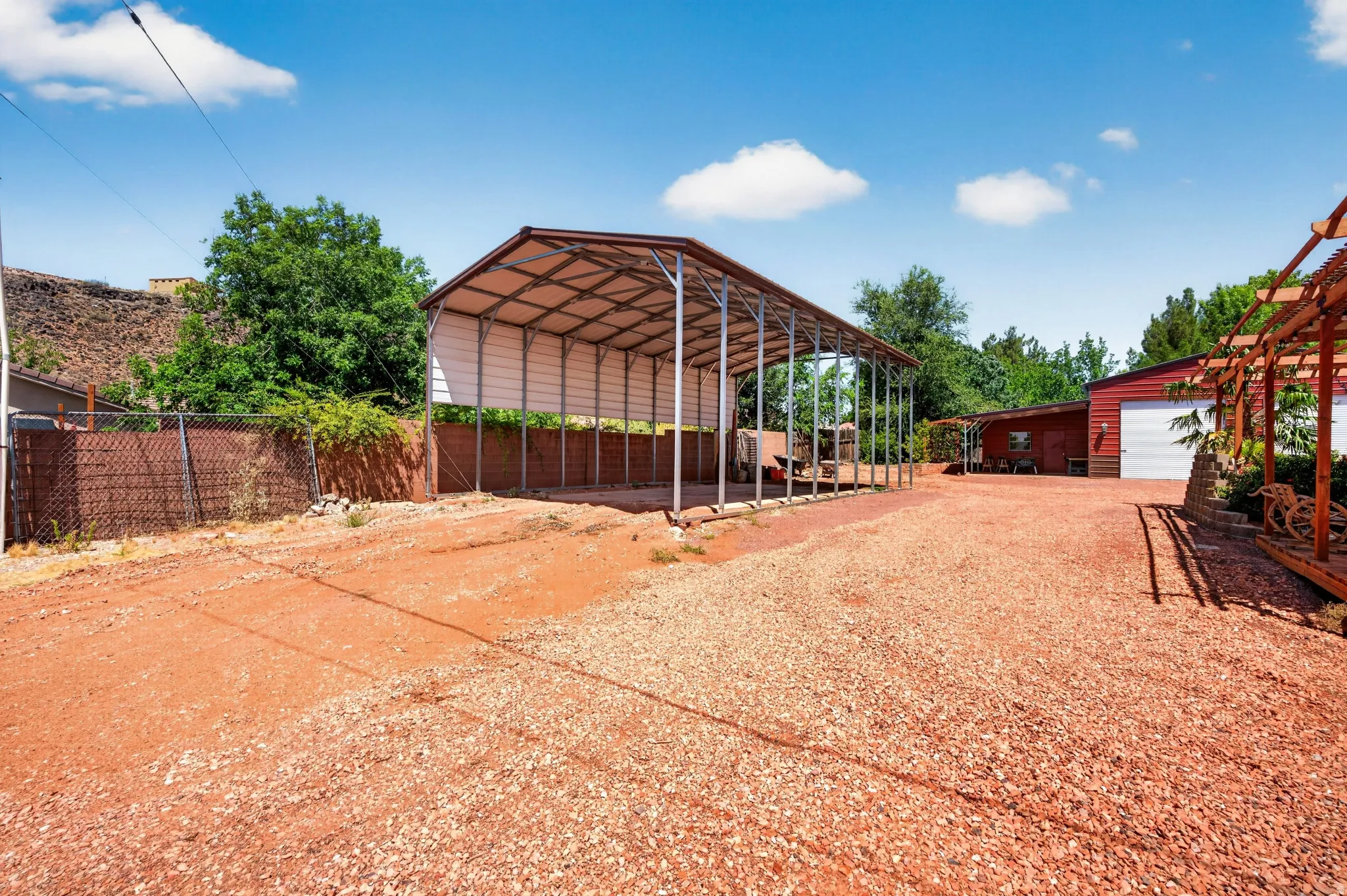 Expansive gravel lot featuring a large covered carport structure, a red outbuilding with a garage door, and a wooden pergola