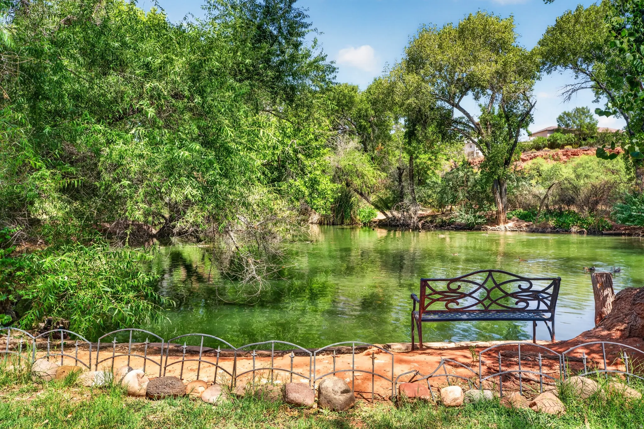 Waterfront property featuring a decorative metal bench, mature trees, natural rock accents, and a low metal fence