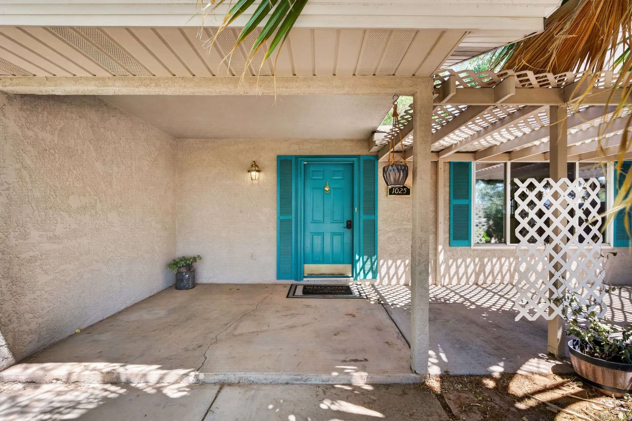Covered front entry featuring a bright turquoise door with coordinating shutters, stucco exterior, and an overhead trellis