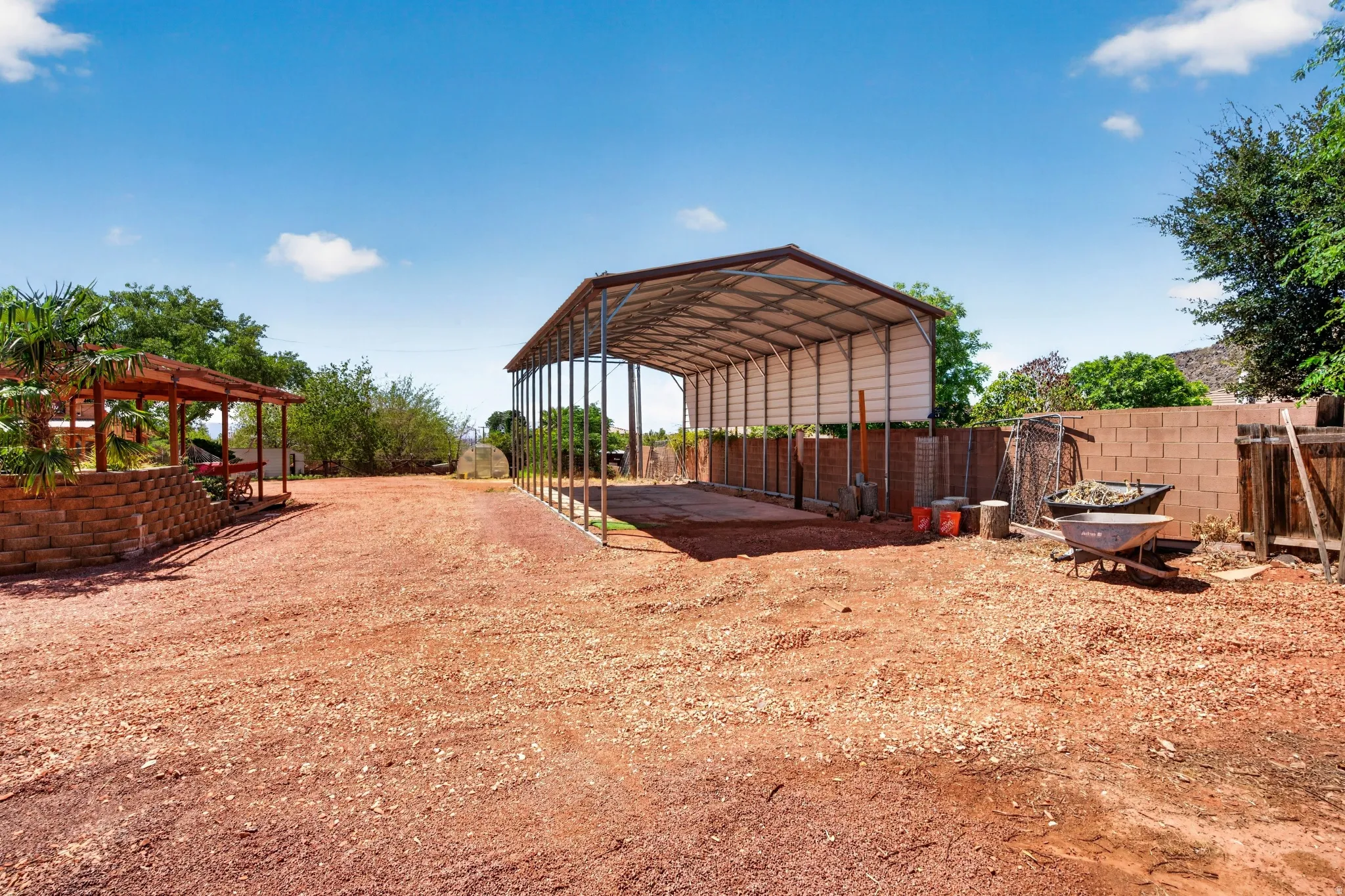 Expansive gravel yard featuring a large covered metal structure, a covered wooden patio with a block retaining wall, and a block privacy fence