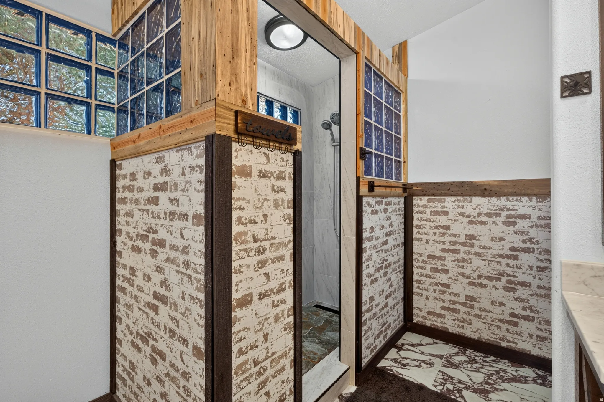 Shower enclosure featuring glass block accents, wood-finish trim, distressed brick-style wall panels, a showerhead, and patterned flooring