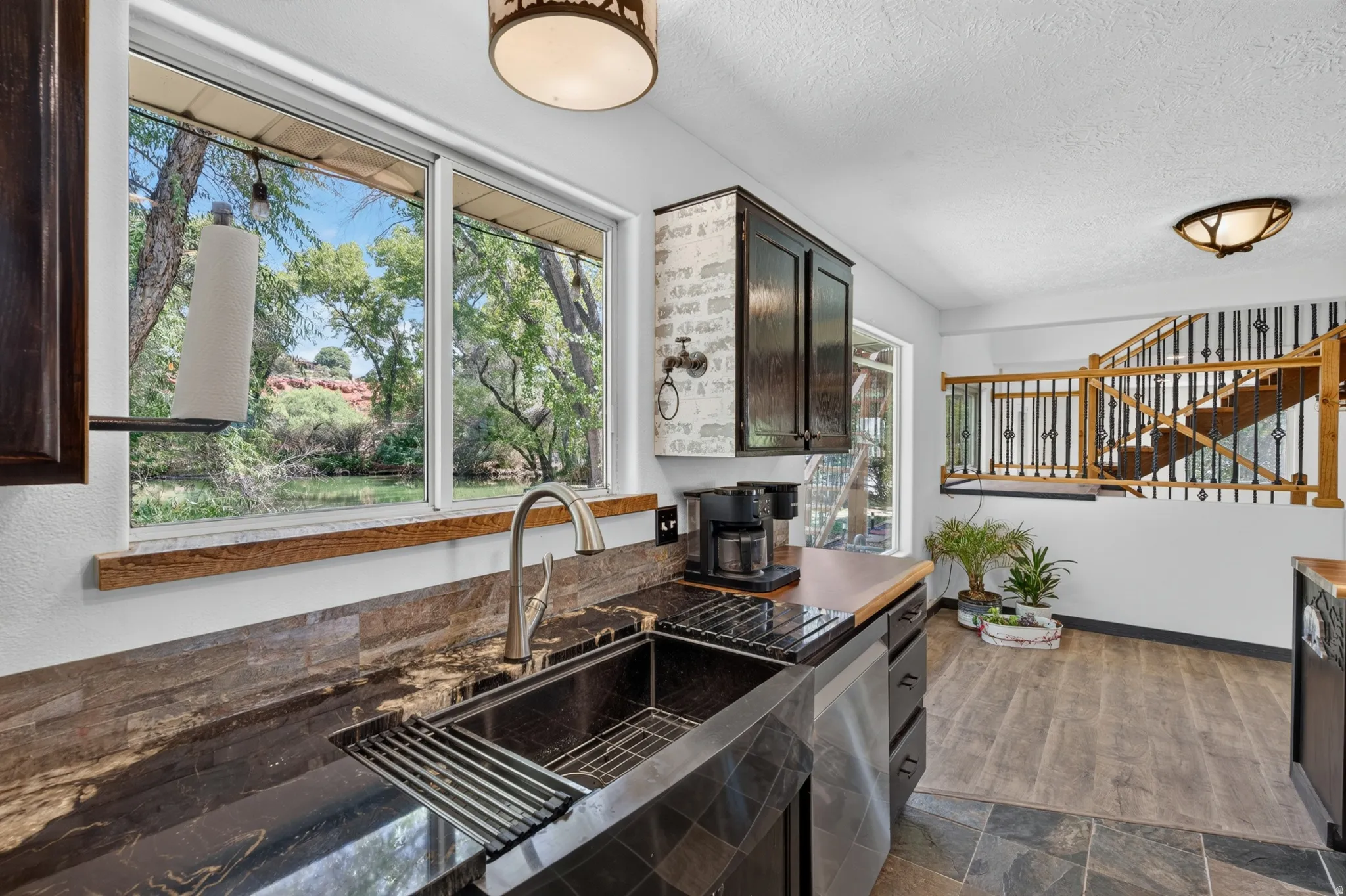 Kitchen area featuring dark cabinetry, a large window overlooking mature trees, and a black workstation sink with a brushed nickel faucet