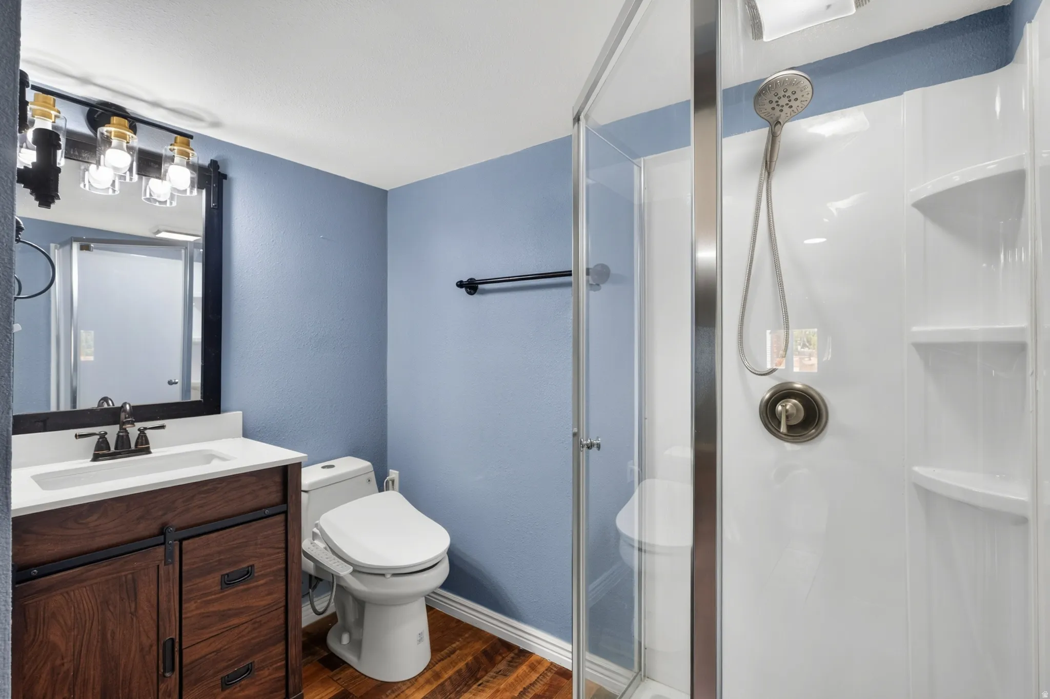 Bathroom featuring a glass-enclosed shower, a vanity with a white countertop and wood-finish cabinetry, a contemporary light fixture, and wood-finish flooring