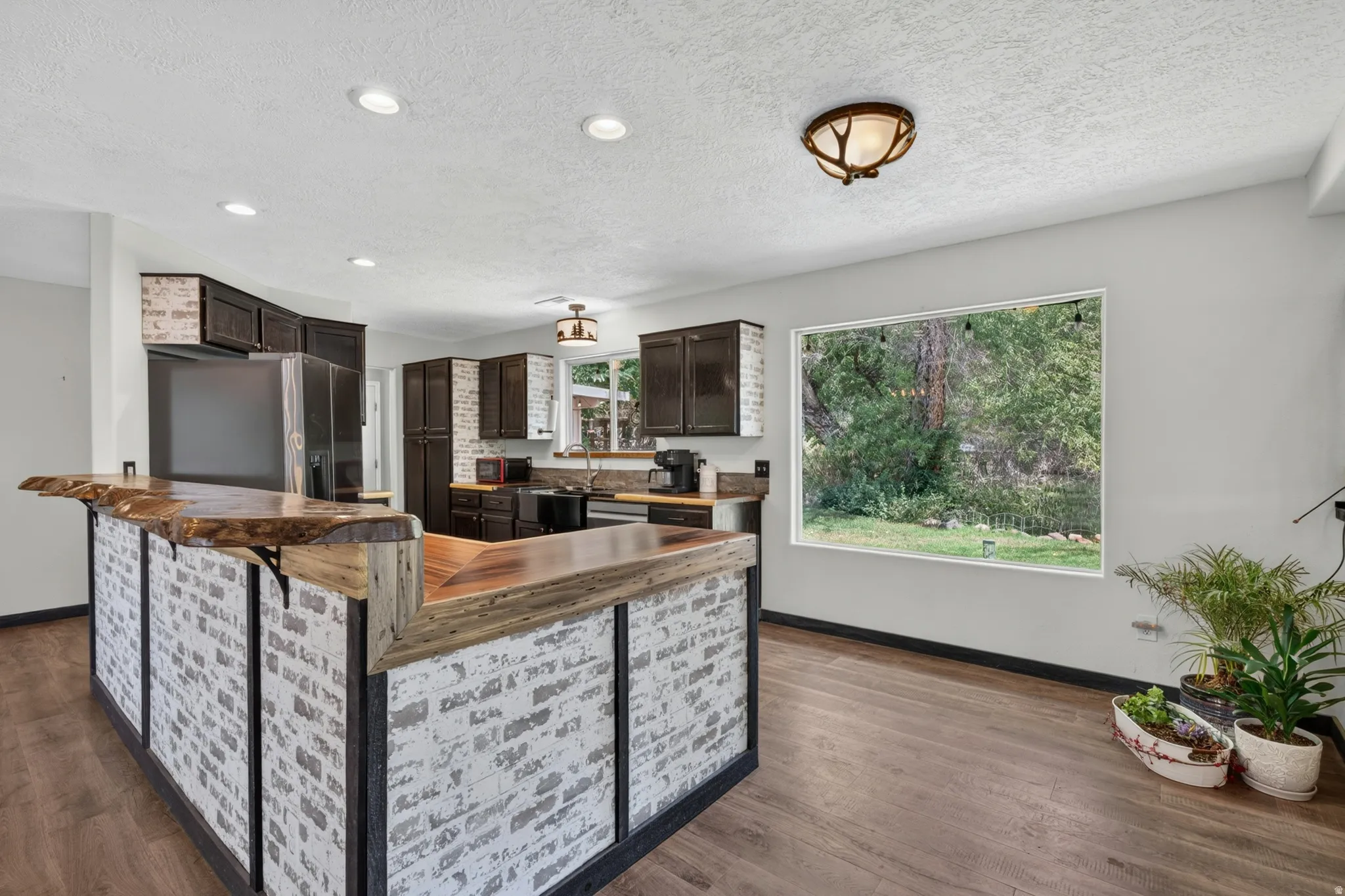 Kitchen featuring a white brick-style island with a live-edge wood countertop, dark wood-finish cabinetry, a large window, and wood-finish flooring