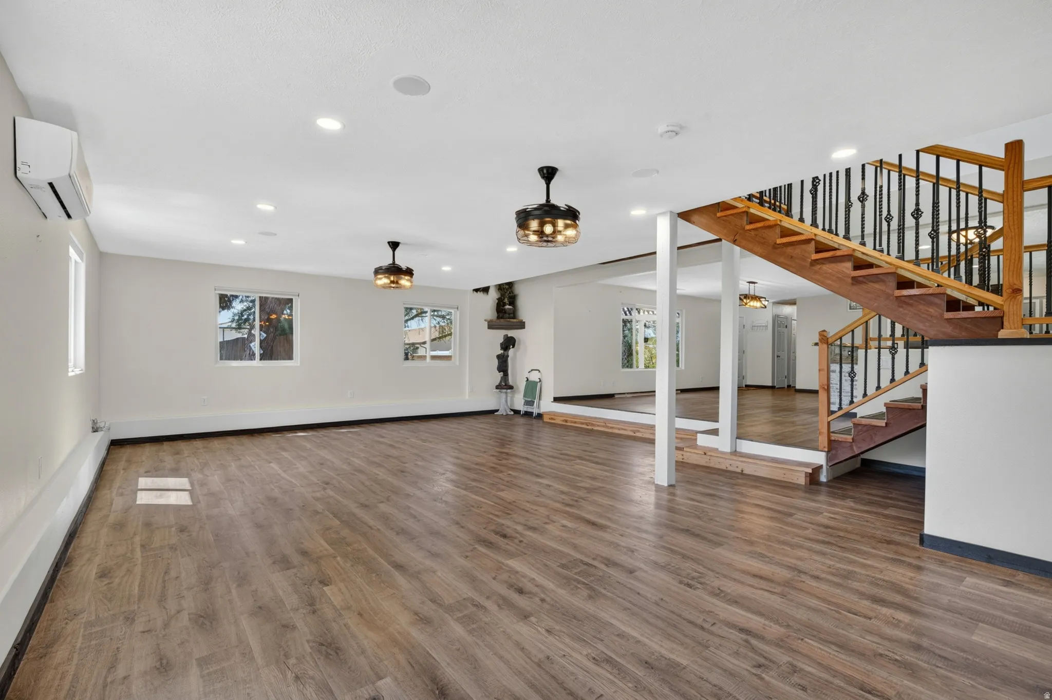 Expansive interior space featuring wood-finish flooring, a prominent wooden staircase with wrought iron balusters, recessed lighting, and multiple windows