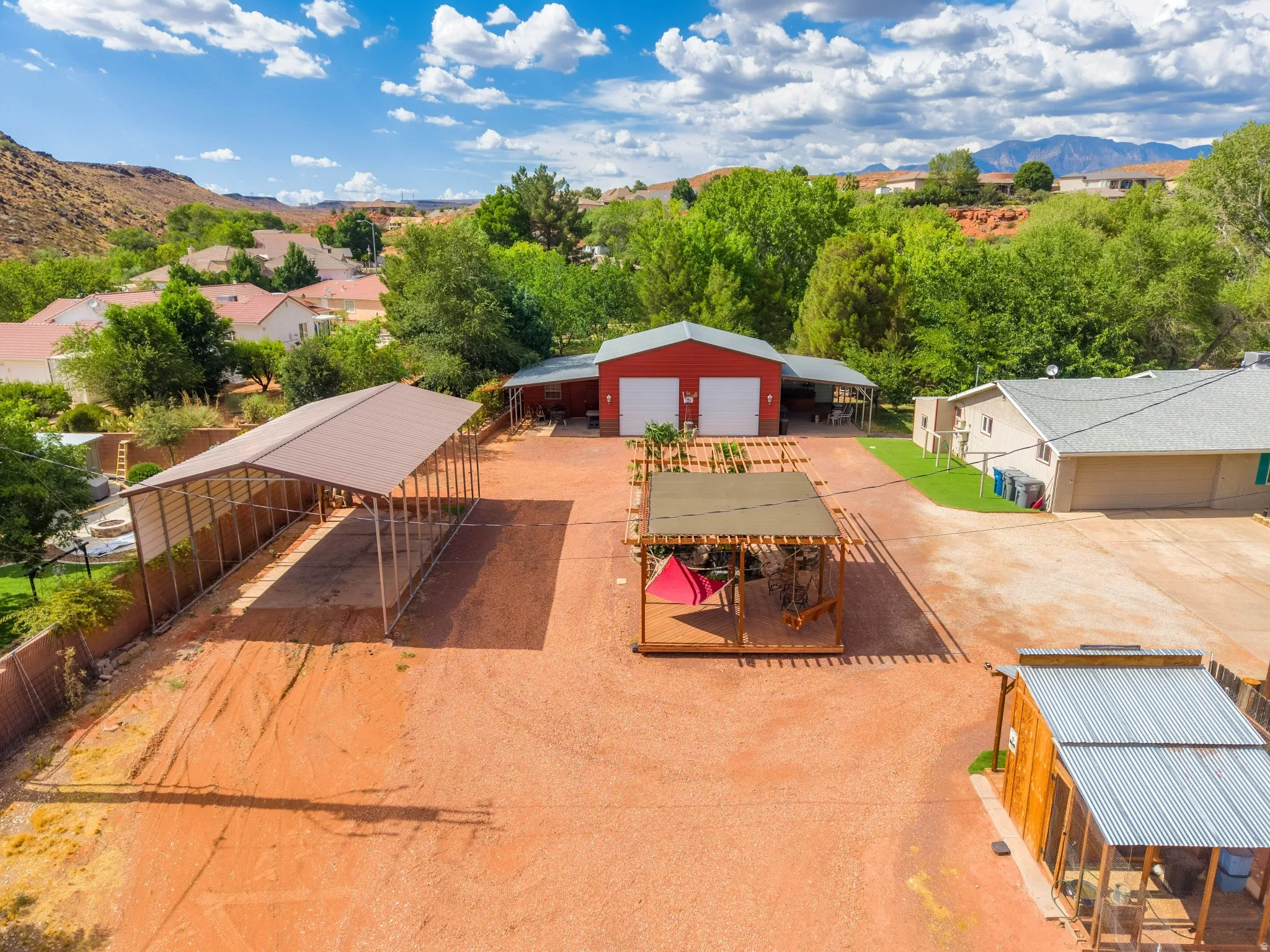 Expansive property featuring a red metal workshop with two garage doors, an extended covered parking structure, a separate single-story residence with an attached garage, and a covered outdoor patio area