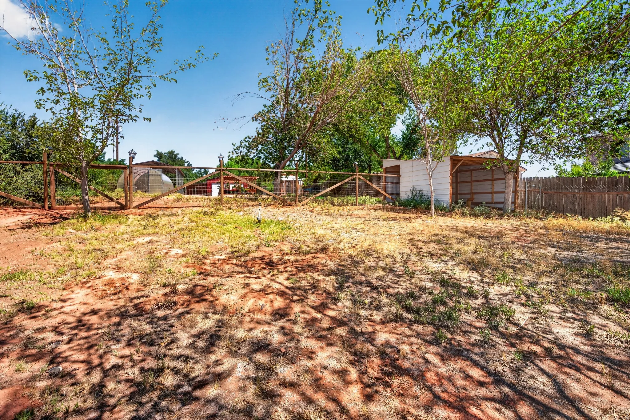 Expansive outdoor area featuring a wooden fence, multiple mature trees, and a distant greenhouse structure