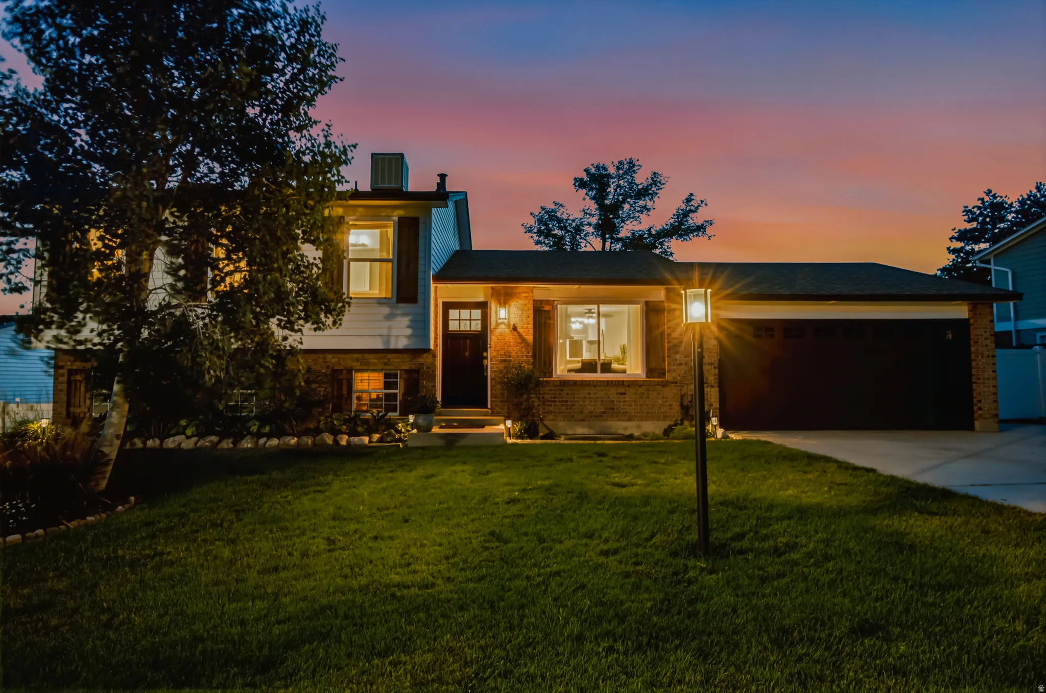 Brick Colonial home illuminated by landscape lighting, featuring a two-car attached garage, manicured lawn with mature trees, and a modern front entrance with illuminated architectural detailing