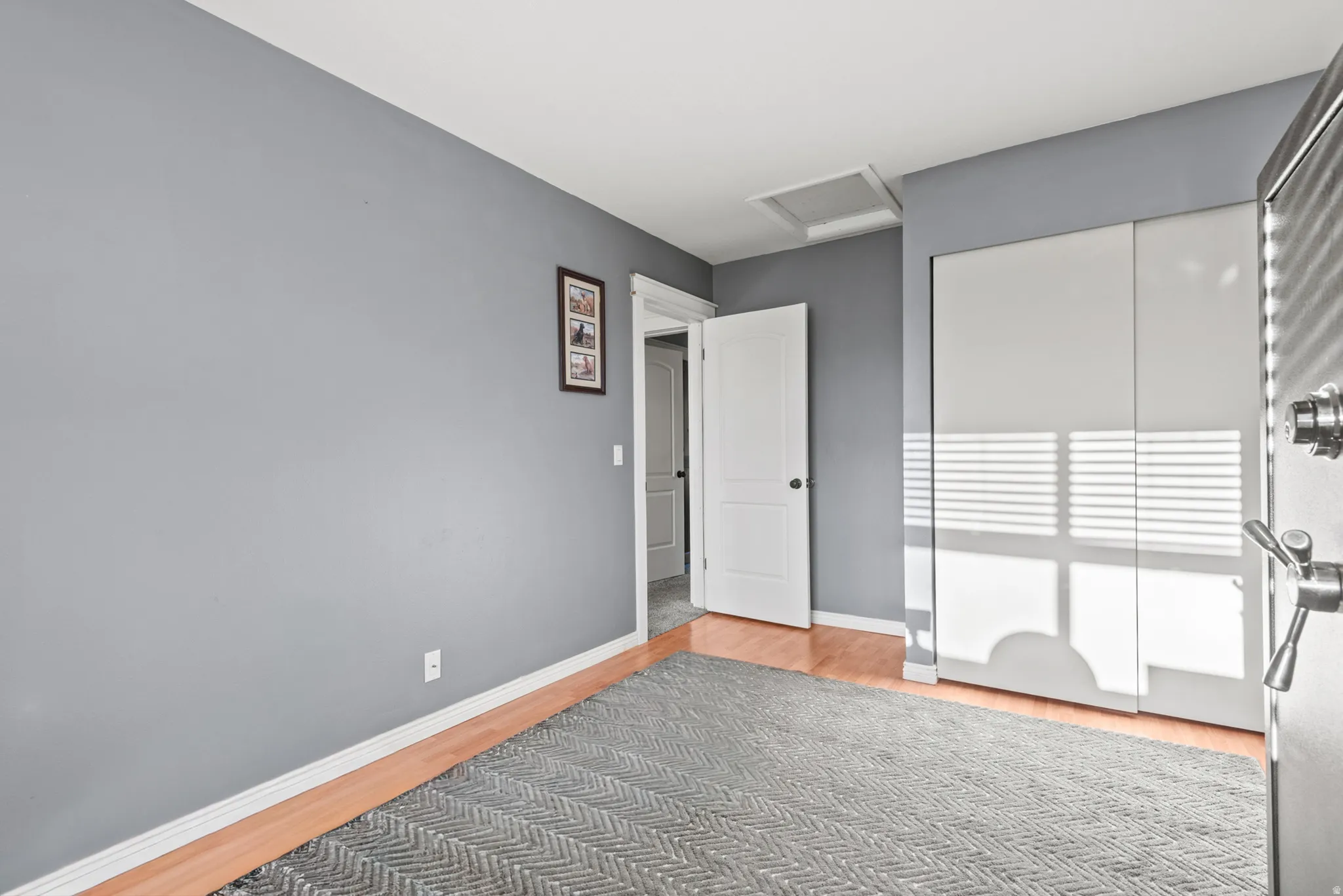 Bright secondary bedroom with warm wood-finish flooring, neutral gray walls, and abundant natural light from sliding glass doors with integrated blinds