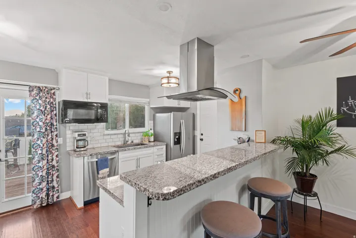 Modern kitchen featuring white cabinetry, stainless steel appliances including a gas range with overhead hood ventilation, granite-style countertops, and a large waterfall-edge island with seating