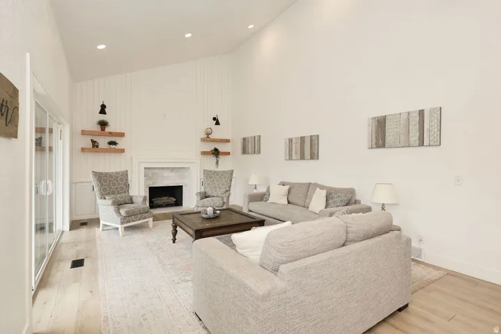 Bright living space featuring vaulted ceilings, light wood-finish flooring, a fireplace with a stone surround and white mantel, built-in shelving, and recessed lighting