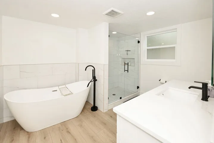 Modern bathroom featuring a freestanding soaking tub with a matte black floor-mounted faucet, a walk-in shower with a glass enclosure and black hardware, a single vanity with a white countertop and integrated sink, and wood-finish flooring