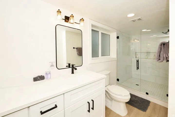 Modern bathroom featuring a single vanity with a white countertop and matte black hardware, a frameless glass shower enclosure with white tile, a window, and wood-finish flooring
