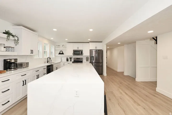 Contemporary kitchen featuring an oversized island with a waterfall countertop, white cabinetry with matte black hardware, stainless steel appliances, and wood-finish flooring