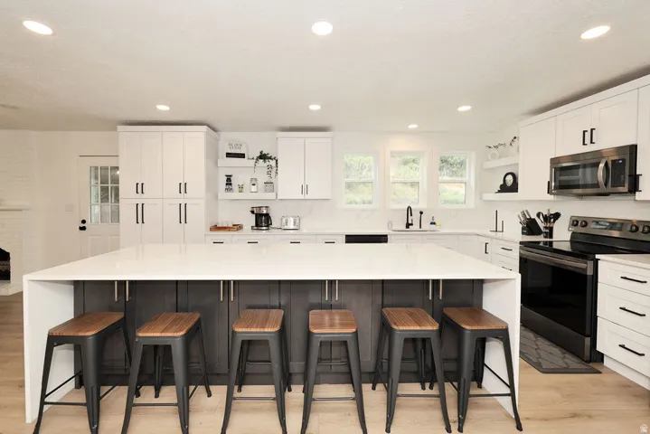 Kitchen featuring a waterfall-edge island with seating, light-tone wood-finish flooring, white upper and lower cabinetry with contrasting dark hardware, stainless steel appliances, and recessed lighting