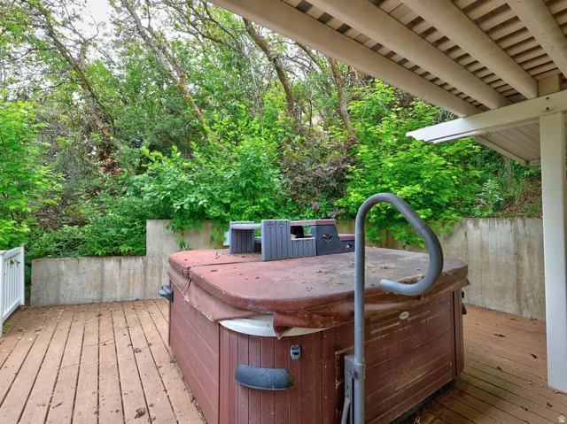 Expansive outdoor deck featuring a hot tub with a safety rail, a white railing, and a slatted patio cover, surrounded by mature trees and natural landscaping