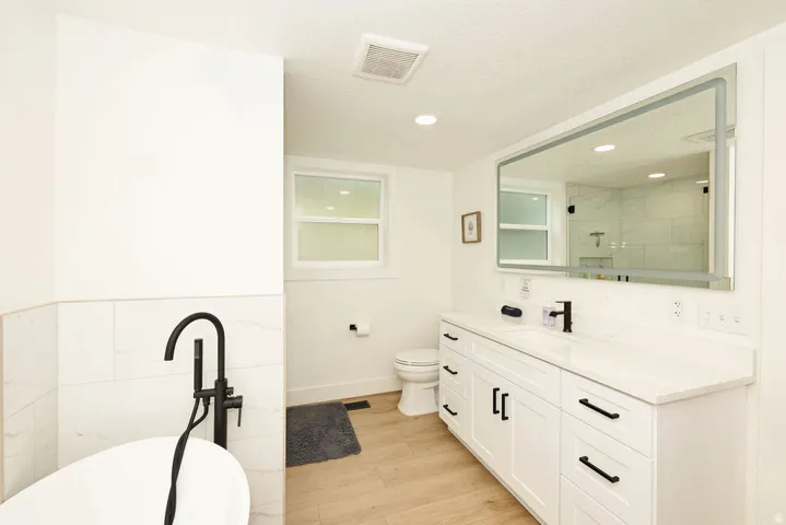 Bathroom featuring a freestanding tub with a matte black floor-mounted filler, a double vanity with white cabinetry, a rectangular illuminated mirror, and wood-finish flooring