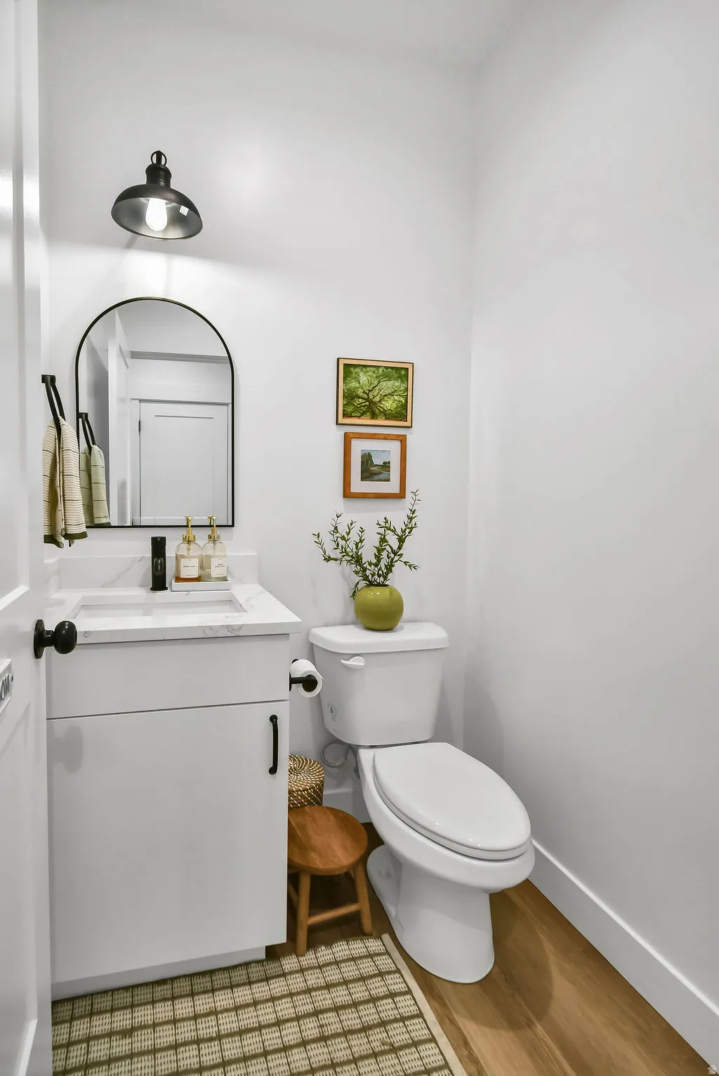 Powder room featuring a white single-sink vanity with a quartz countertop, a black arched mirror, and wood-finish flooring