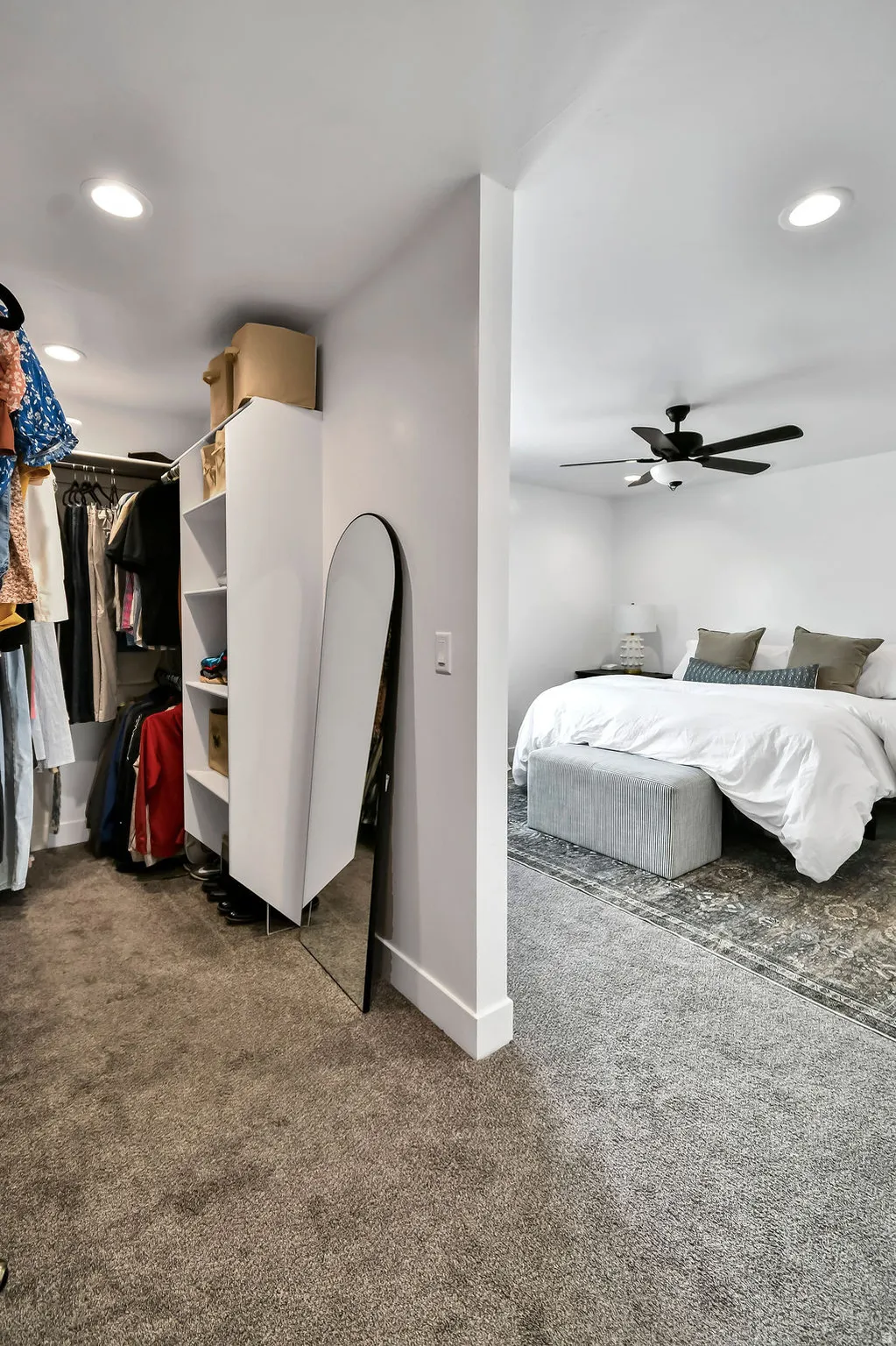 Carpeted walk-in closet with built-in shelving, recessed lighting, and a partial view into an adjacent carpeted room with a ceiling fan