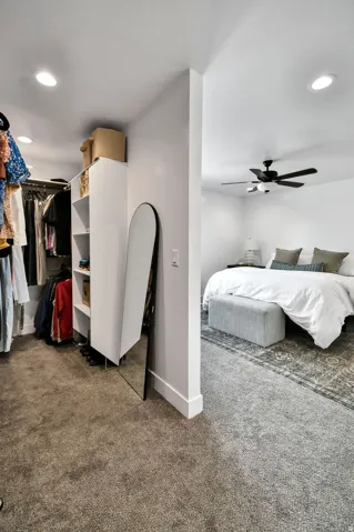 Carpeted walk-in closet with built-in shelving, recessed lighting, and a partial view into an adjacent carpeted room with a ceiling fan