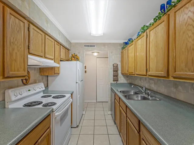 Galley kitchen featuring wood-finish cabinetry, white appliances, a dual-basin stainless steel sink, and light-toned tile flooring
