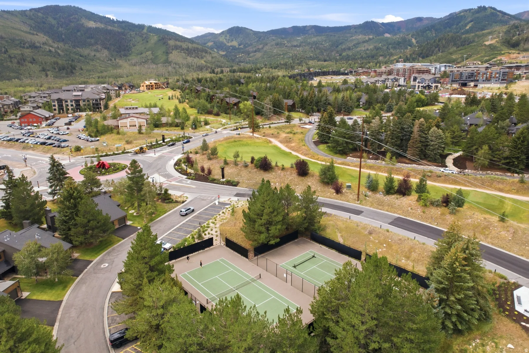 Aerial perspective featuring two outdoor tennis courts surrounded by mature trees, paved roadways, and a mountainous backdrop