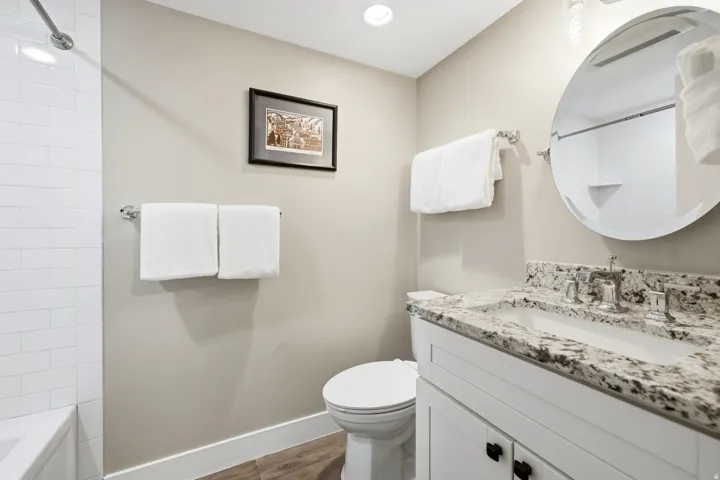 Bathroom featuring a white vanity with a speckled countertop, a round mirror, and a white subway tile shower surround