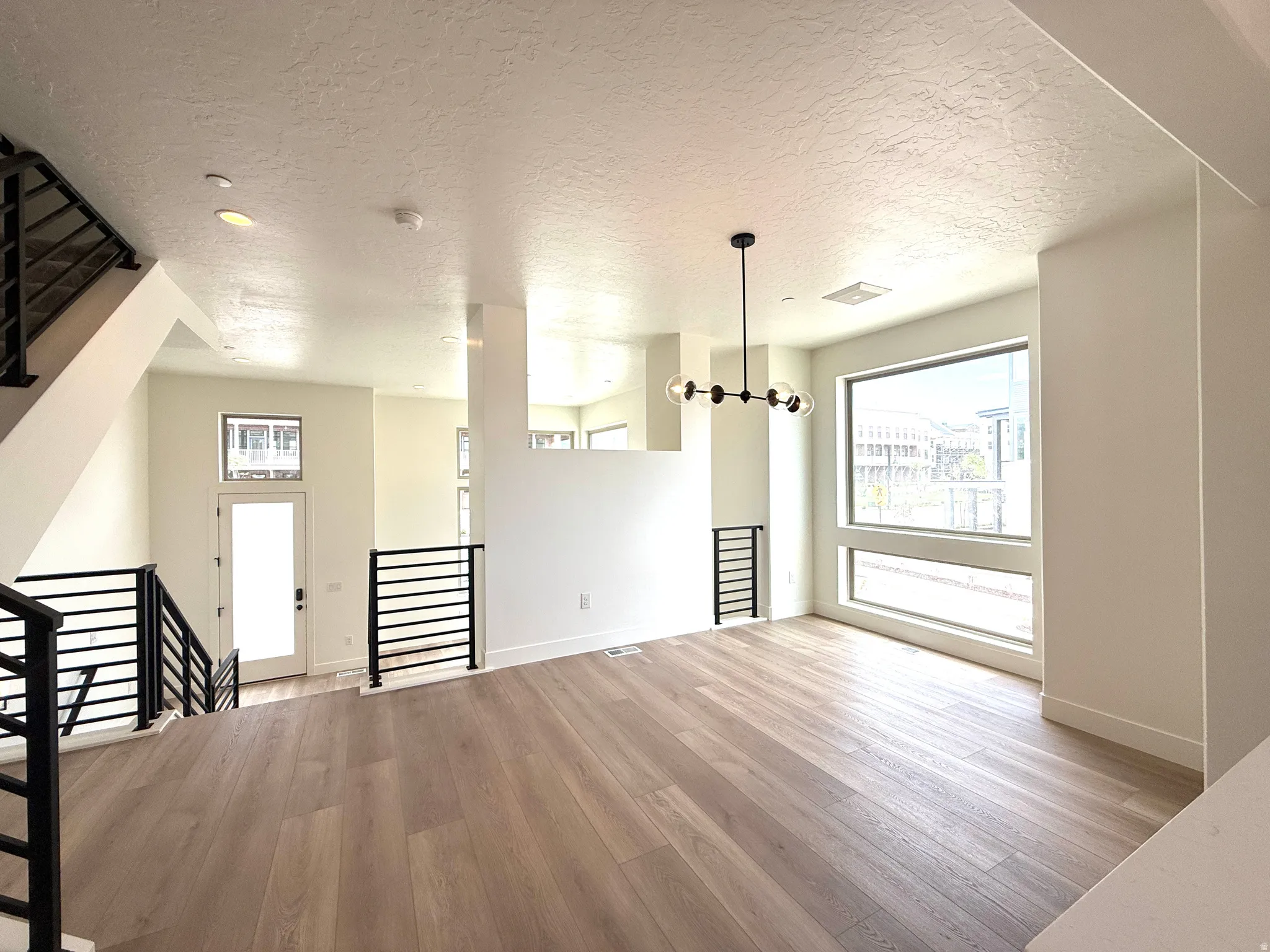 Open-concept living space featuring wood-finish flooring, a contemporary chandelier, large picture windows, and black metal railings on the staircase and balcony