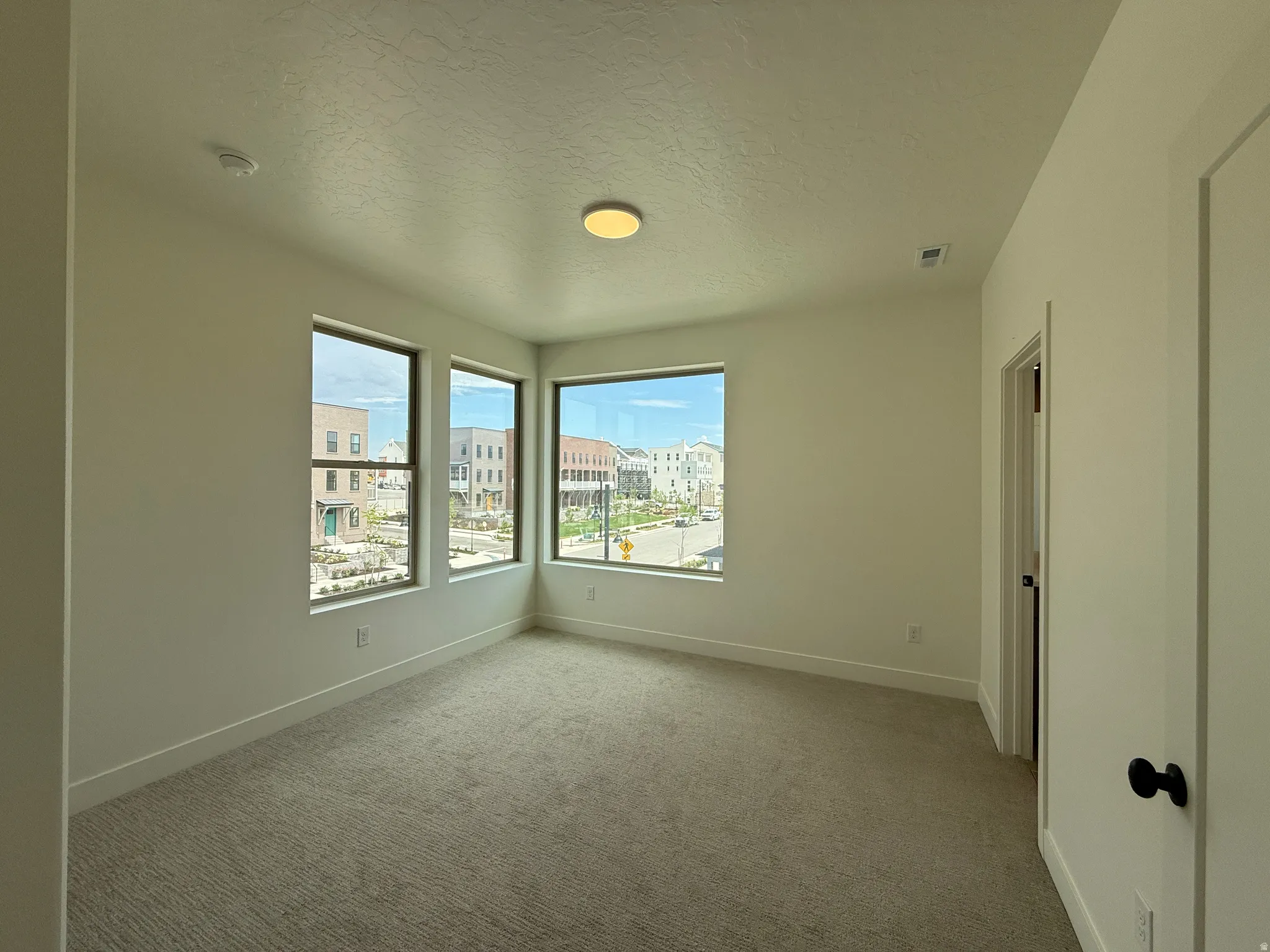 Corner room featuring three large windows, neutral-toned carpet, white walls, and a flush-mount ceiling light fixture