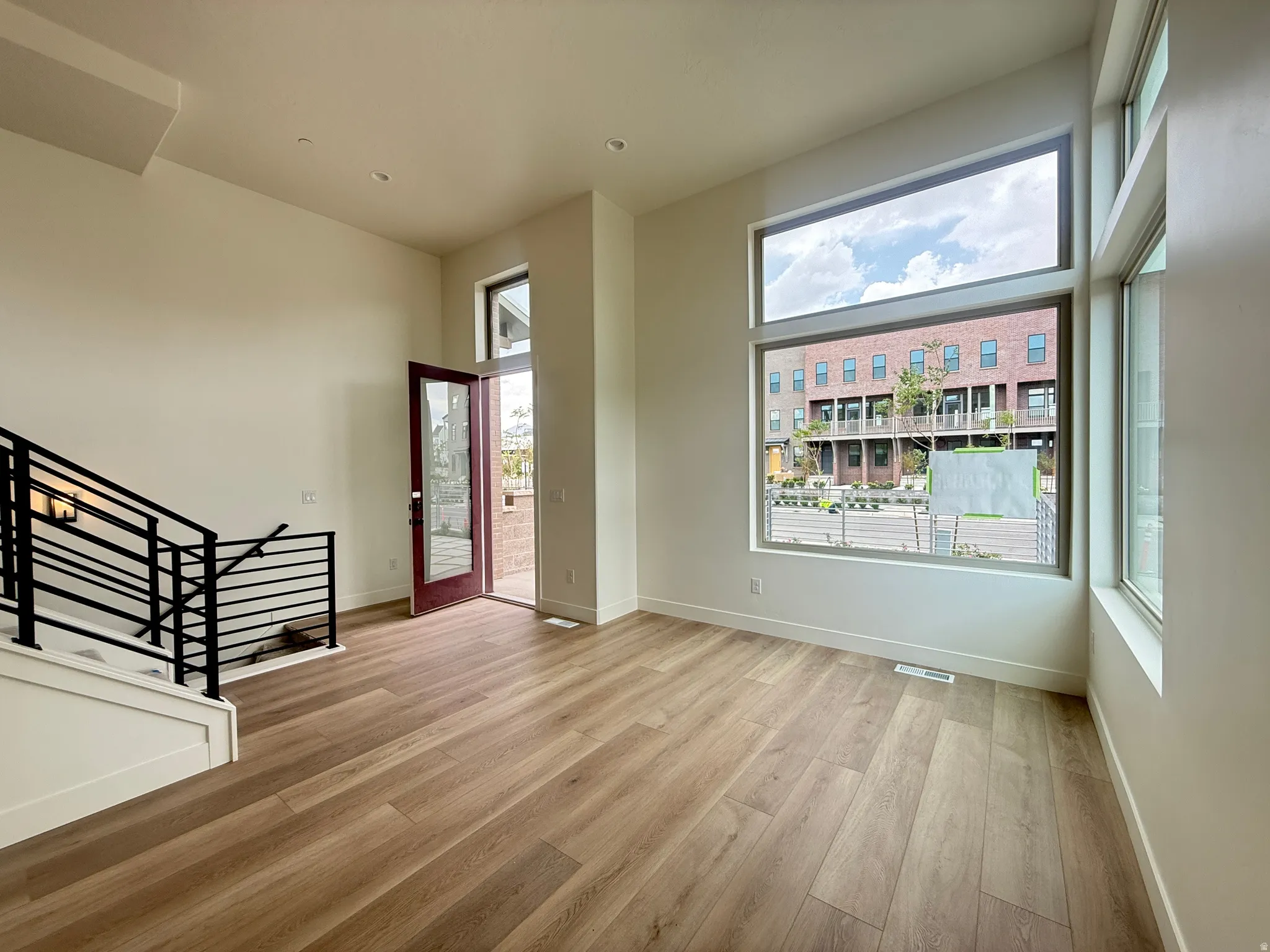 Spacious open-concept room featuring wide-plank wood-finish flooring, a contemporary black metal staircase railing, and large windows with transom panels