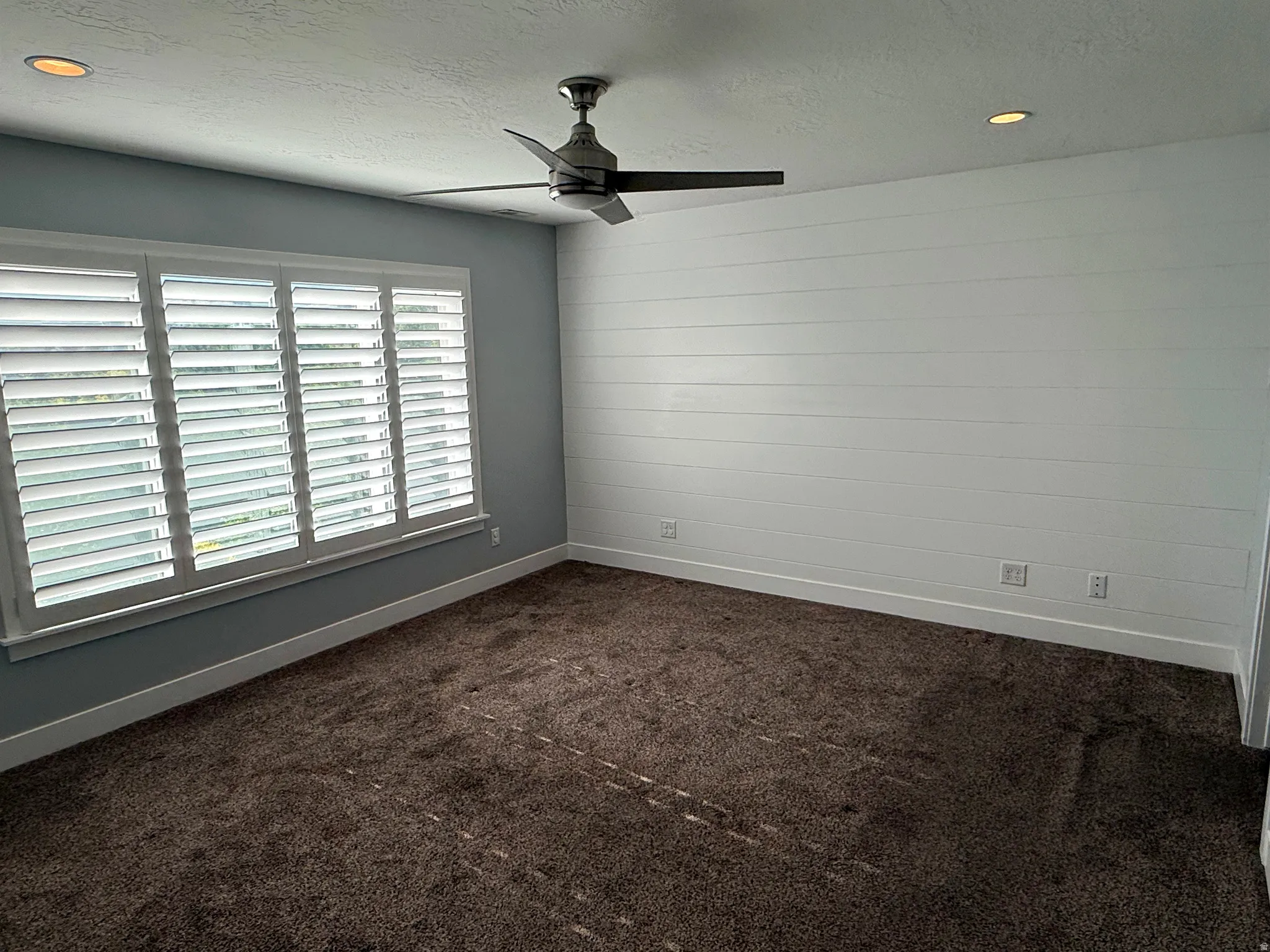 Spacious room featuring a white shiplap accent wall, white plantation shutters, a modern ceiling fan, recessed lighting, and soft brown carpet