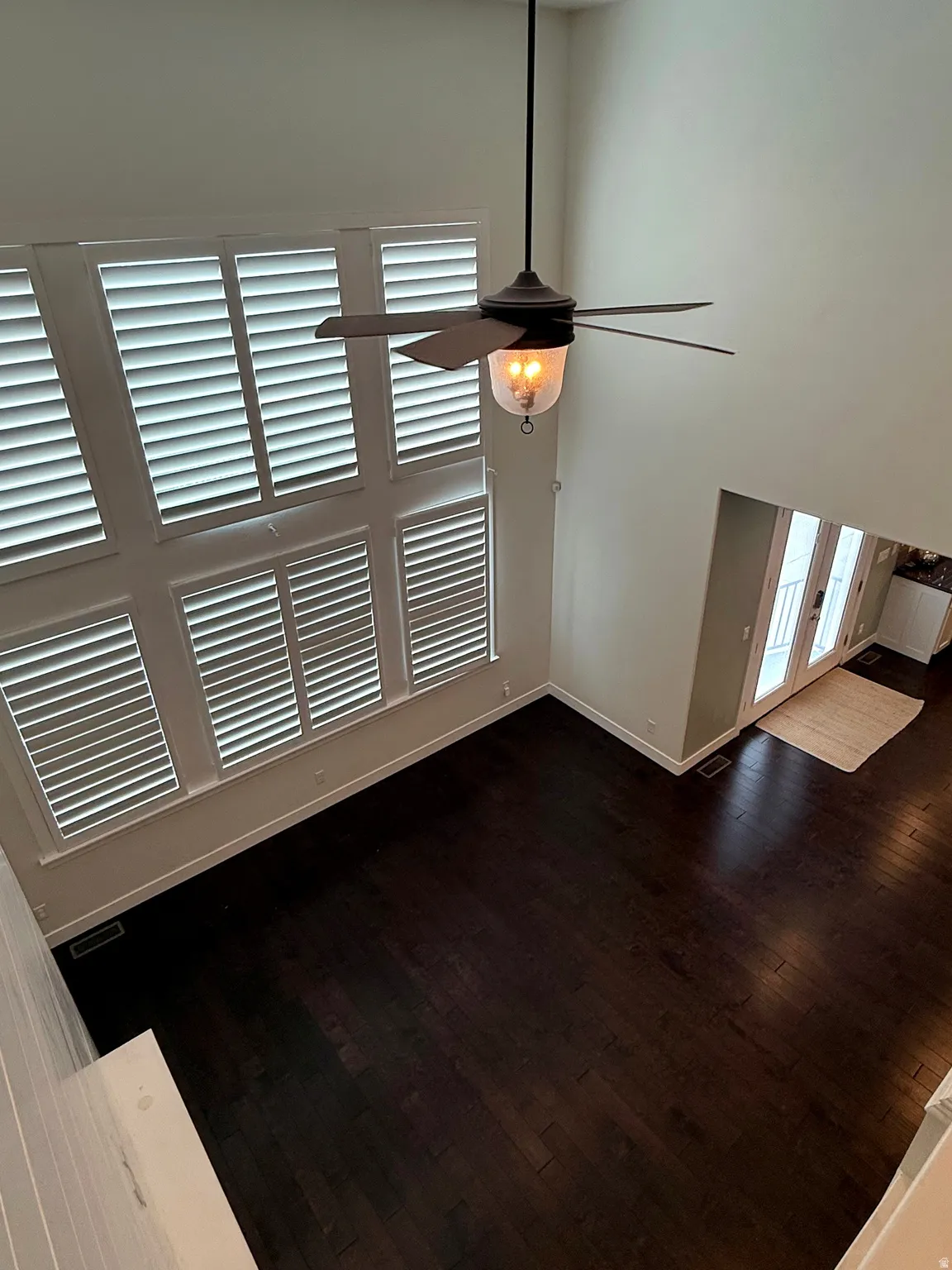 Two-story room featuring dark wood-finish flooring, a contemporary ceiling fan with integrated lighting, and multiple windows with plantation shutters
