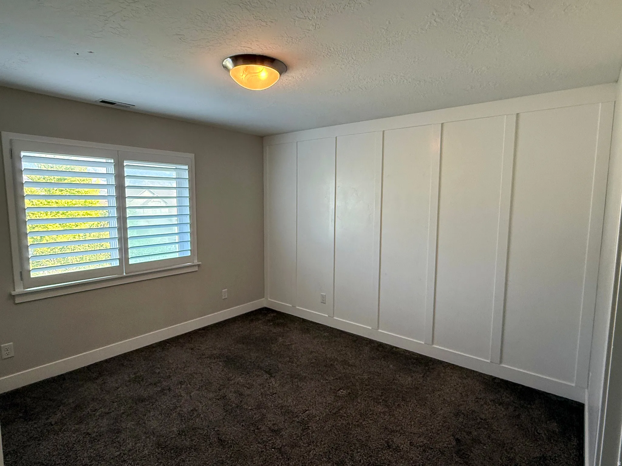 Interior room featuring a board and batten accent wall, window with white plantation shutters, and dark carpet flooring