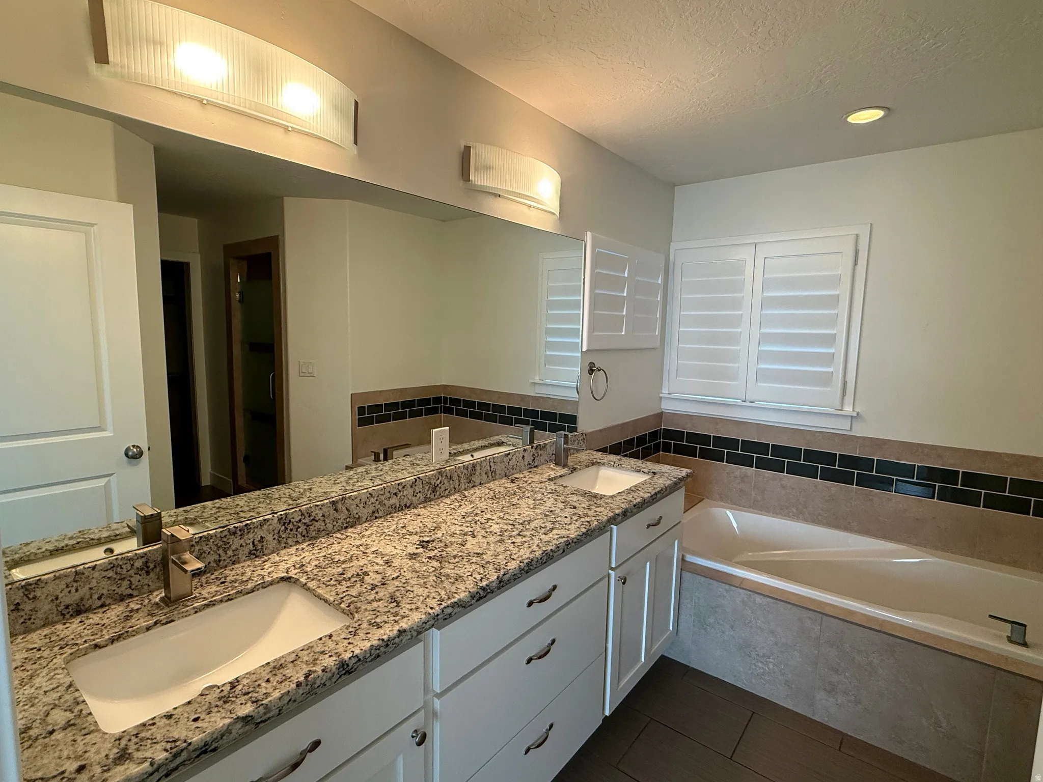 Bathroom featuring a dual vanity with granite countertops, built-in bathtub, tile flooring, and white plantation shutters