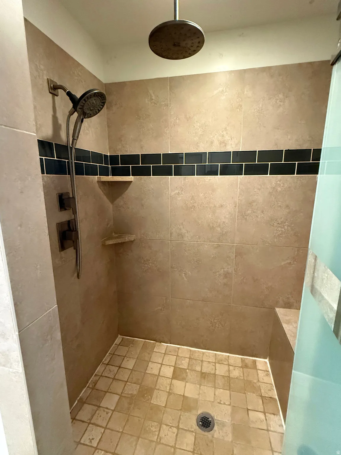 Walk-in shower with large format tan wall tiling, featuring a black accent tile stripe, dual showerheads including a rain showerhead, a built-in bench, and corner shelving